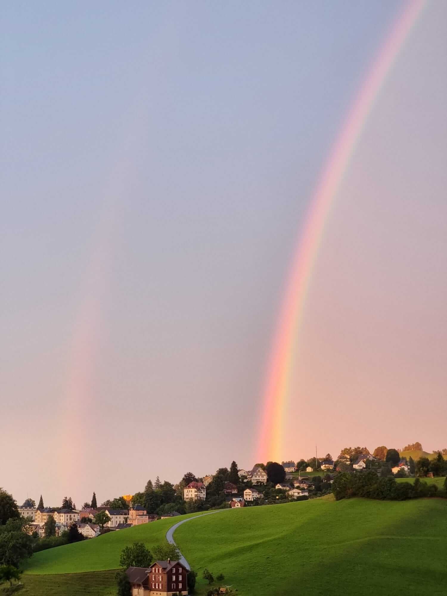 Ein leuchtender Regenbogen spannt sich über einen pastellfarbenen Himmel über einem grünen Hügel mit verstreuten Häusern und Bäumen in der Ostschweiz. Ein schwacher zweiter Regenbogen ist sichtbar, und eine kurvenreiche Straße führt zu einem kleinen Haus - ein Symbol für Abschied nehmen und Neuanfang.