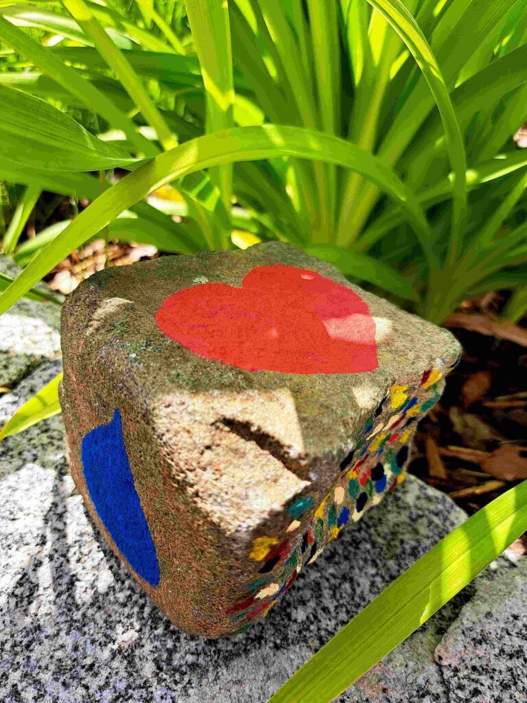 Ein bemalter Felsen mit einem roten Herz, einer blauen Form und bunten Punkten sitzt auf einer Steinfläche, umgeben von leuchtend grünen Blättern - ein sanftes Symbol, das in der Trauerbegleitung in der ganzen Ostschweiz häufig zu sehen ist.