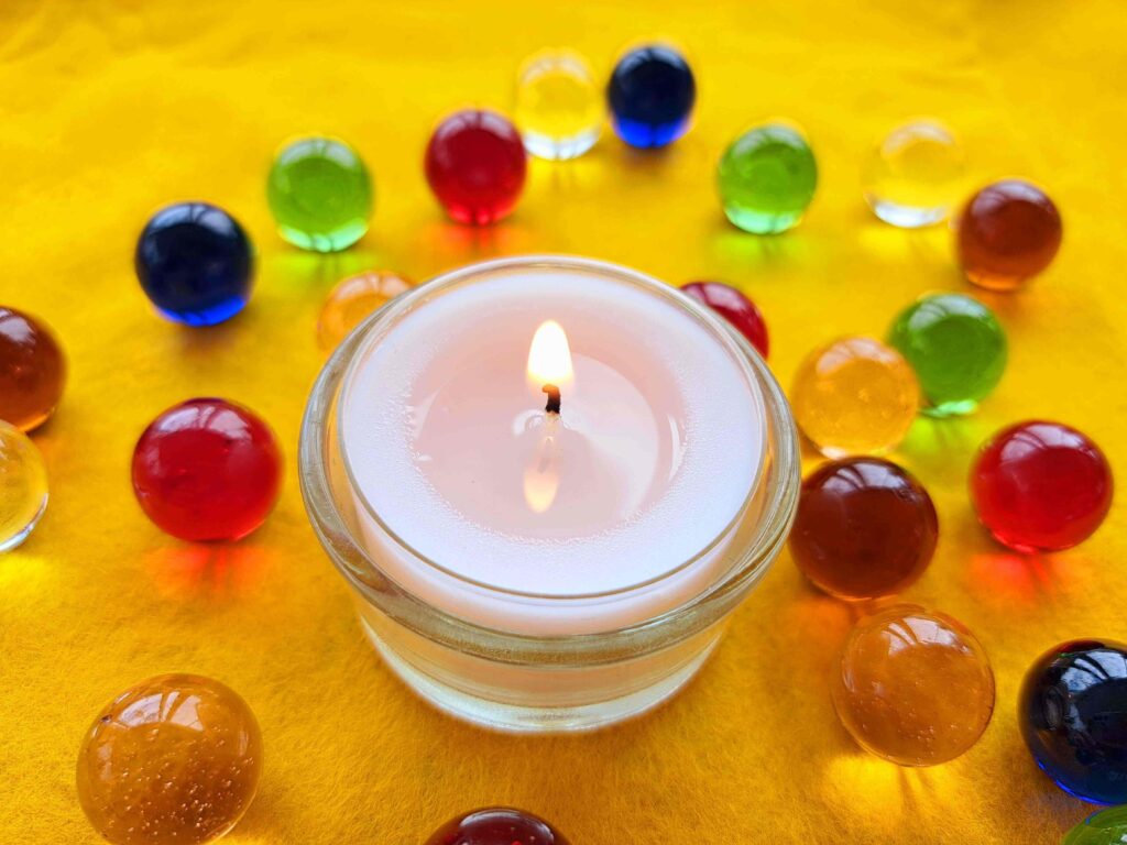 Eine brennende weiße Kerze in einem Glashalter ist umgeben von bunten Glasmurmeln auf einer leuchtend gelben Oberfläche, die an die sanften Trauerwellen des Abschieds erinnern - ein Moment der stillen Besinnung, der an Ostschweizer Traditionen erinnert.