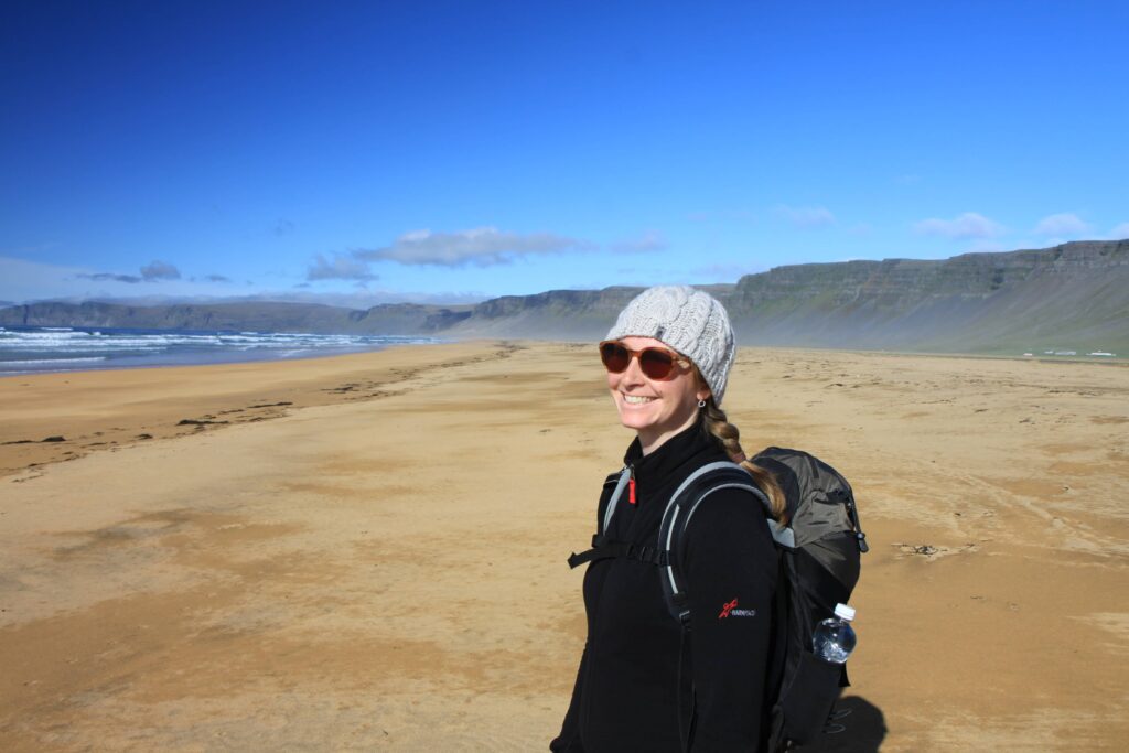 Eine Frau mit Sonnenbrille, Strickmütze und Rucksack steht lächelnd an einem weiten, leeren Sandstrand mit Wellen wie Trauerwellen und fernen Felsen unter strahlend blauem Himmel in der Ostschweiz.