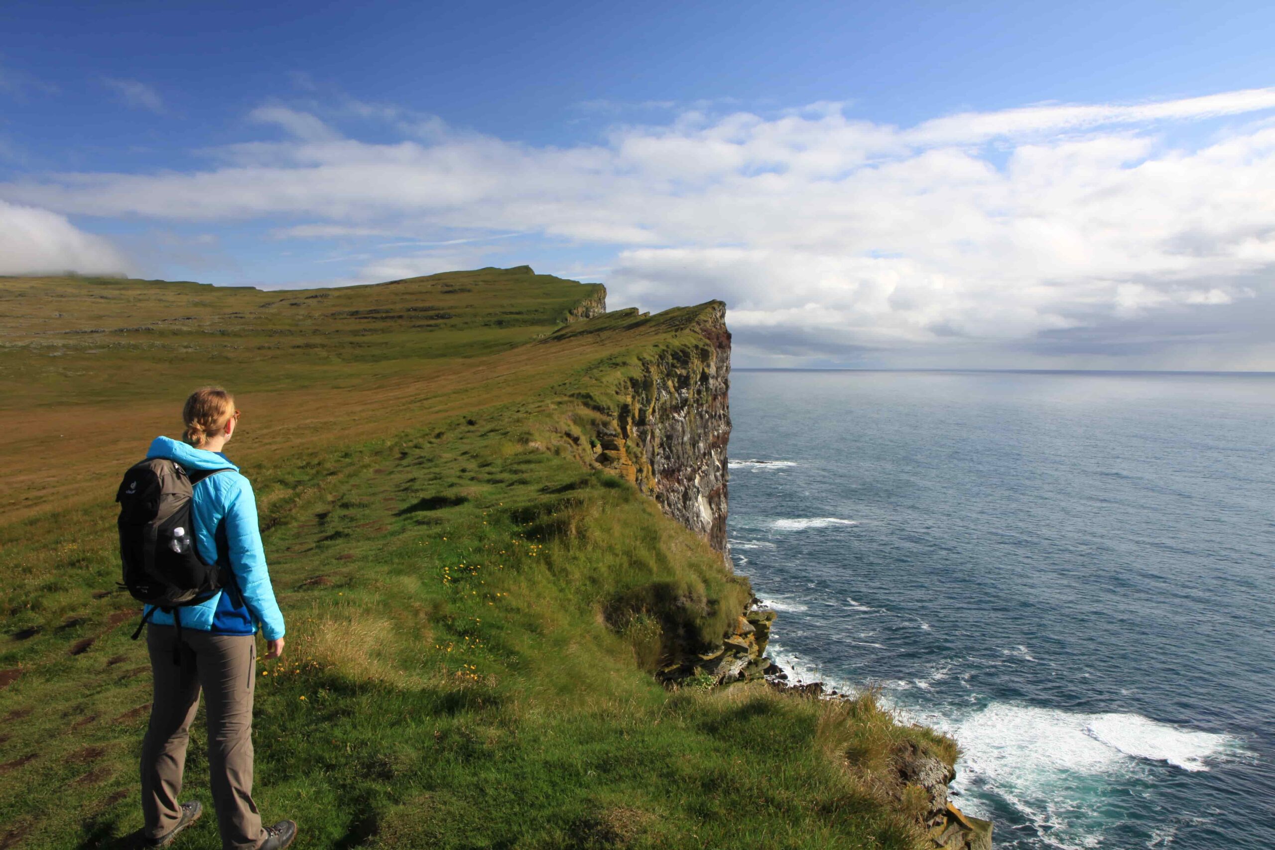 Eine Person mit blauer Jacke und Rucksack steht am Rande einer grasbewachsenen Klippe in der Ostschweiz und blickt auf das Meer mit seinen Wellen und dem bewölkten Himmel darüber.