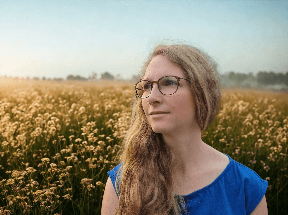 Eine Frau mit langem, gewelltem blondem Haar und Brille steht in einem Feld mit hohem Gras und Wildblumen, trägt ein blaues Oberteil und blickt nachdenklich in die Ferne unter einem klaren Ostschweizer Himmel.