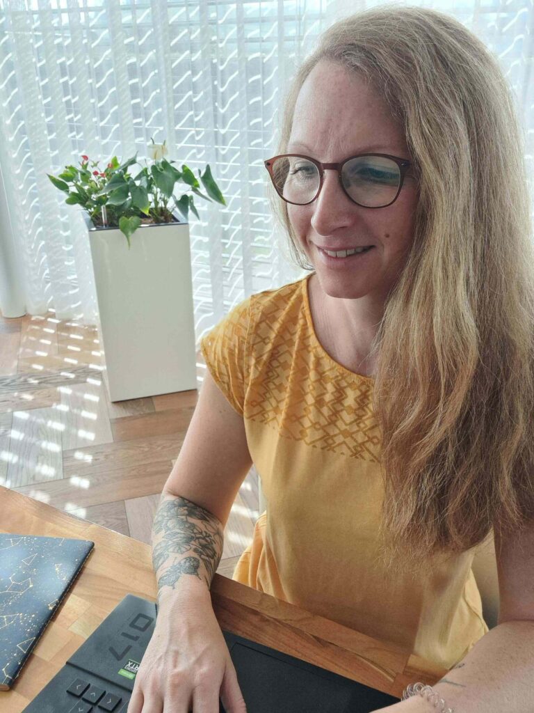 Eine Frau mit langen blonden Haaren und Brille, die ein gelbes Oberteil trägt, sitzt an einem Schreibtisch und benutzt einen Laptop. Während das Sonnenlicht durch die transparenten Vorhänge im Hintergrund fällt, denkt sie über Trauerwellen und den Prozess des Abschiednehmens nach. Eine Topfpflanze und ein Notizbuch liegen in der Nähe.