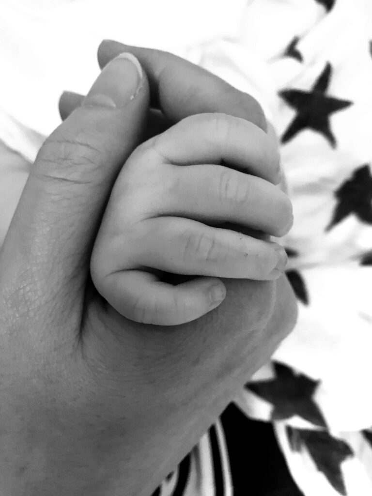 Ein Schwarz-Weiß-Foto in Nahaufnahme, das die Hand eines kleinen Kindes zeigt, die sanft den Finger eines Erwachsenen hält, mit einer sterngemusterten Decke im Hintergrund - es fängt einen ruhigen Moment des Abschiednehmens und die Zärtlichkeit der Trauerbegleitung ein.