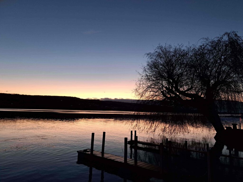Silhouette eines Baumes und eines hölzernen Stegs an einem ruhigen See bei Sonnenuntergang in der Ostschweiz, mit einer dunklen Uferlinie und einem von Orange zu Tiefblau übergehenden Himmel - ein Bild, das an Frieden und stille Besinnung erinnert, ideal für Trauerbegleitung.