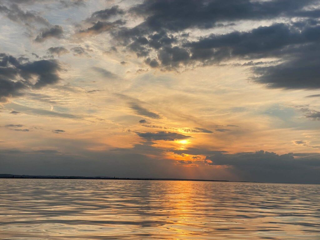 In einem ruhigen Gewässer in der Ostschweiz spiegeln sich die sanften Orange- und Gelbtöne eines Sonnenuntergangs wider. Der Himmel ist wolkenverhangen und die Sonne ist teilweise am Horizont verborgen - sanfte Trauerwellen sind die Folge.