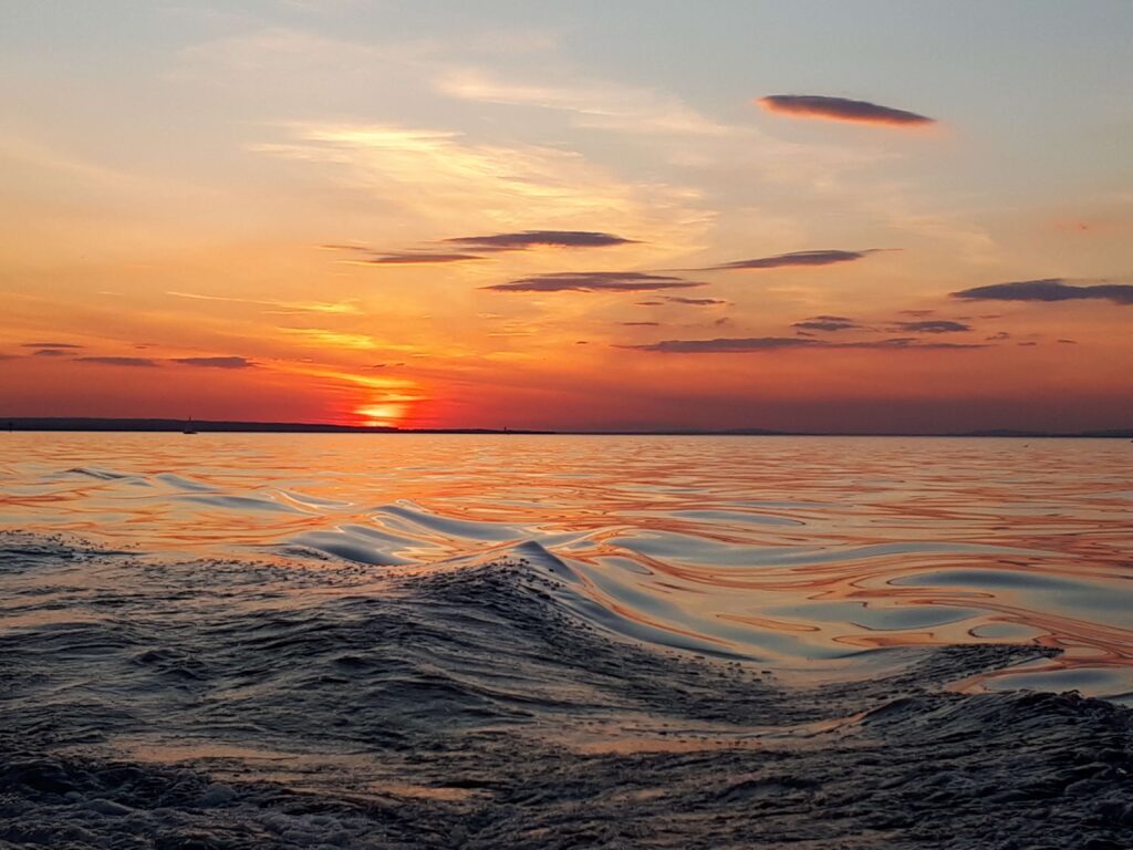 Die Sonne geht über einem ruhigen Meer unter und wirft orange- und rosafarbene Farbtöne über den Himmel und das Wasser, mit sanften Wellen im Vordergrund, die an Trauerwellen erinnern, während man unter den verstreuten Wolken darüber Trost im Abschied nehmen findet.