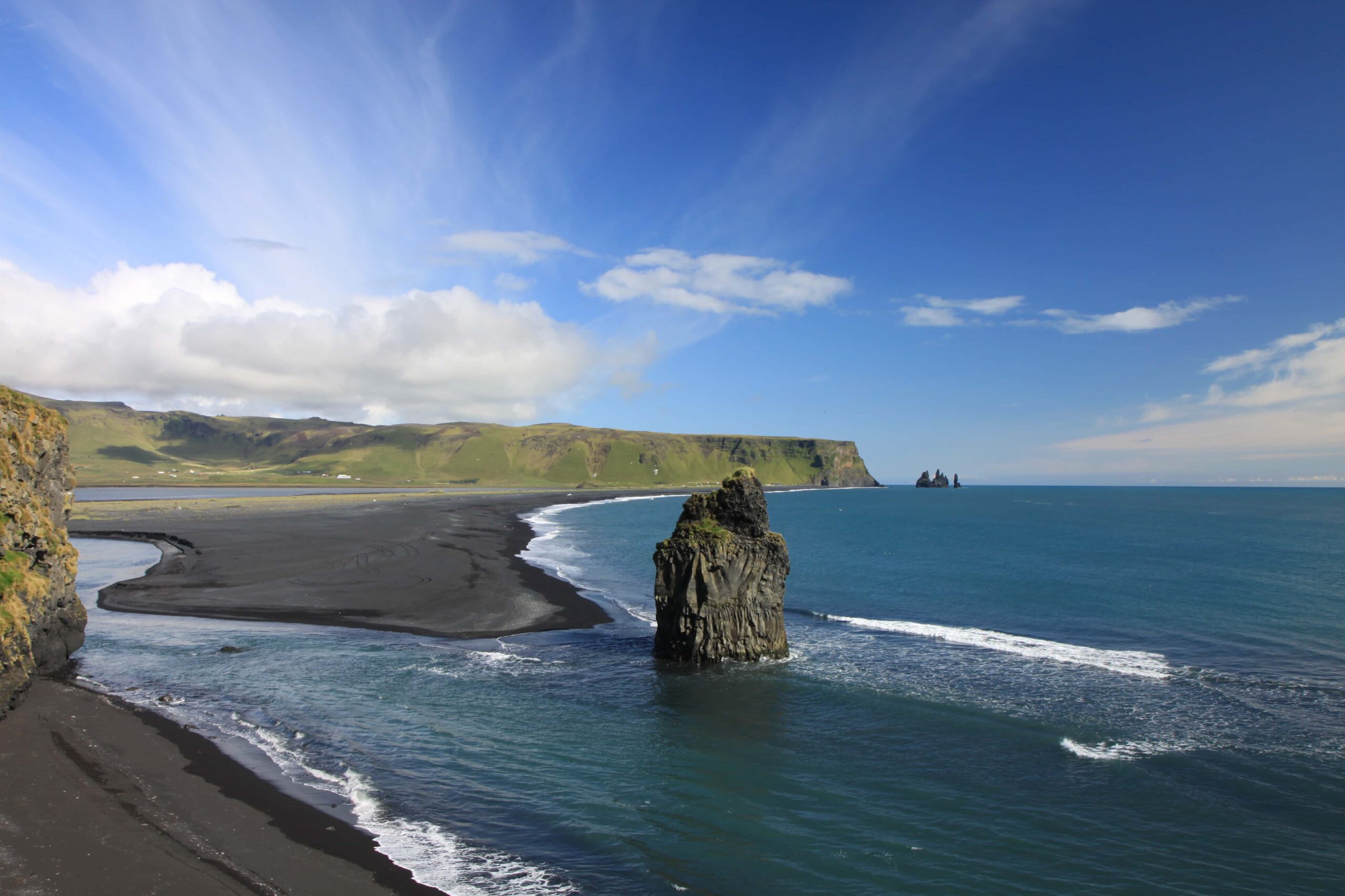 In der Nähe eines schwarzen Sandstrandes in der Ostschweiz ragt ein hoher Seestapel aus dem blauen Meer, mit sanften Wellen und entfernten grünen Klippen unter einem teilweise bewölkten blauen Himmel.