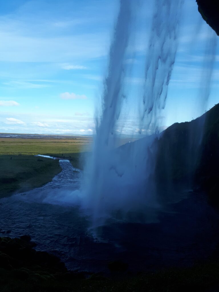 Ein Blick auf einen hohen Wasserfall, der über eine Klippe stürzt, mit Wassernebel und einem Fluss, der durch eine Grasebene unter einem teilweise bewölkten blauen Himmel fließt, erinnert an Trauerwellen in der ruhigen Landschaft der Ostschweiz.