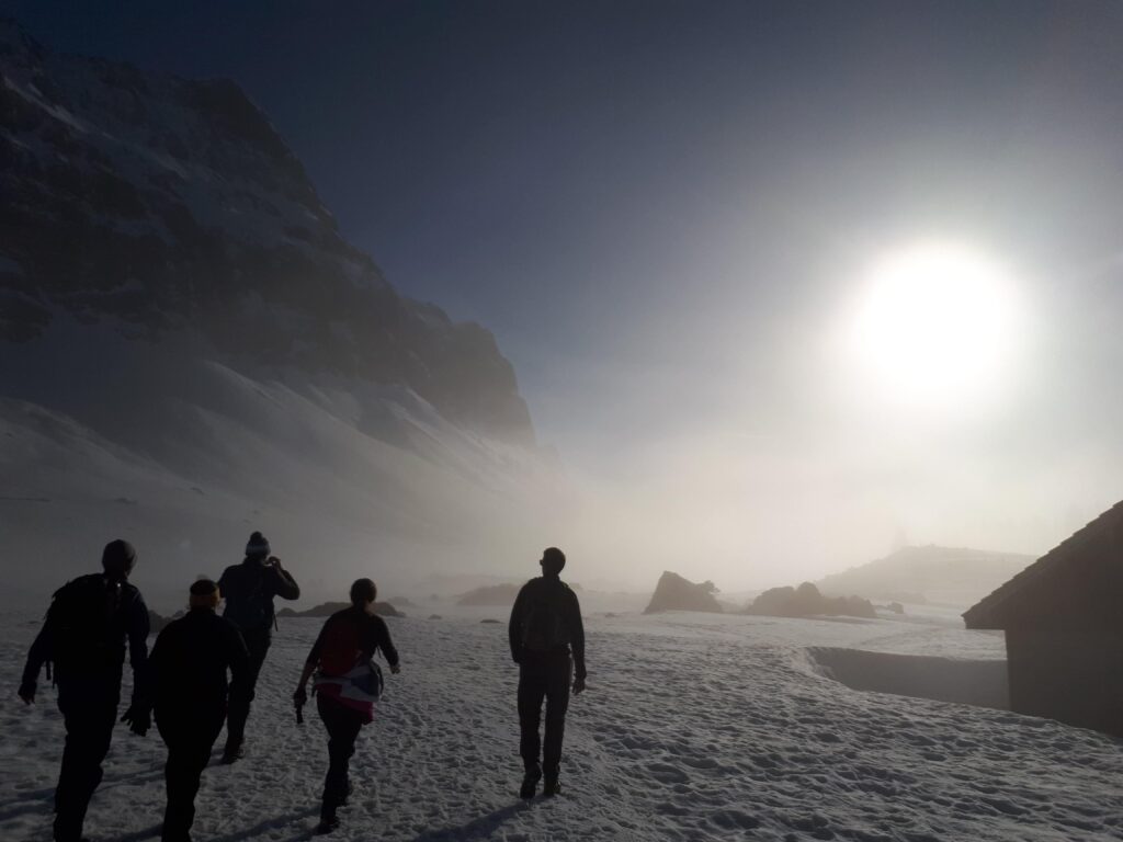 Eine Gruppe von Menschen geht durch eine verschneite Landschaft auf eine strahlende Sonne zu, mit Bergen und einer Hütte, die teilweise durch den Nebel im Hintergrund sichtbar ist - eine stimmungsvolle Szene von Abschied nehmen und sanfter Trauerbegleitung in der Umarmung der Natur.