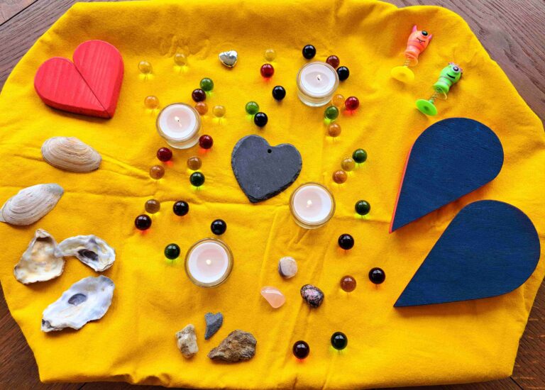 Auf einem gelben Tuch sind Teelichtkerzen, bunte Murmeln, Muscheln, Steine und symbolische Formen in einem dekorativen Muster angeordnet, das die sanfte Unterstützung der Trauerbegleitung und den beruhigenden Rhythmus der Trauerwellen in der Ostschweiz widerspiegelt.