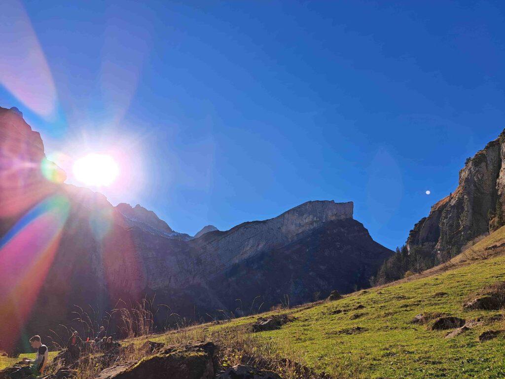 Die Sonne scheint hell über felsige Berge und einen grasbewachsenen Abhang und wirft Lichtreflexe und tiefe Schatten. In der unteren linken Ecke ist eine Person teilweise durch hohes Gras verdeckt - vielleicht nimmt sie leise Abschied, während sanfte Trauerwellen unter dem klaren blauen Himmel vorbeiziehen.