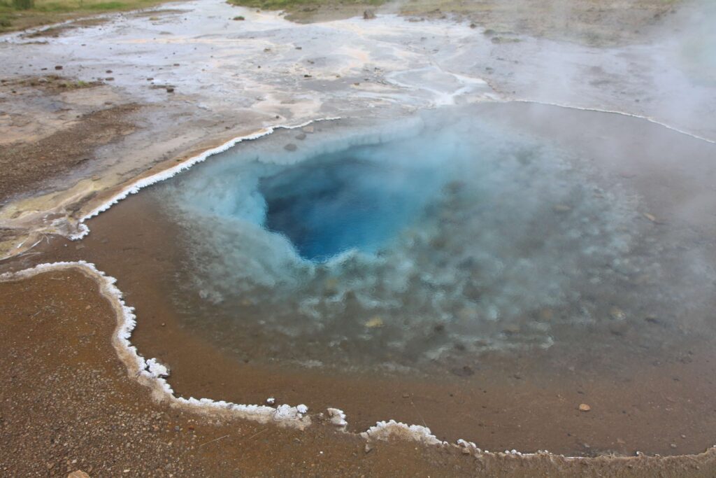 Eine dampfende geothermische Quelle mit leuchtend blauem Wasser, umgeben von mineralischen Ablagerungen und brauner Erde unter einem bedeckten Himmel, erinnert an die stimmungsvollen Trauerwellen, die man oft in den Landschaften der Ostschweiz findet.