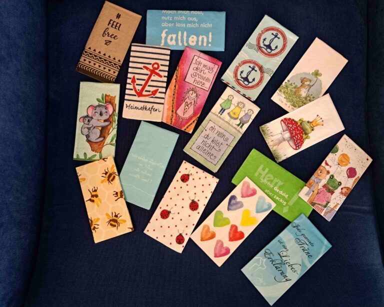Eine Sammlung von farbenfrohen, illustrierten Karten mit verschiedenen Motiven - Tiere, Herzen, Anker, Marienkäfer und deutscher Text - auf einem dunkelblauen Stoffhintergrund, perfekt zum Versenden von Abschiedswünschen in der Ostschweiz.