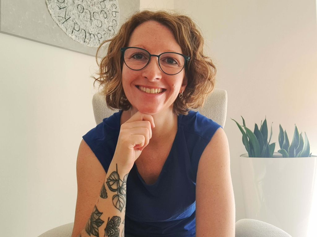 Eine Frau mit kurzem, gewelltem blondem Haar und Brille sitzt in einem weissen Sessel mit einem Bogenhanf im hohen, weissen Blumentopf und einem silbernen Bild auf dem Geduld und Stolz zu lesen sind im Hintergrund, sie trägt ein blaues Oberteil und blickt lächelnd in die Kamera.